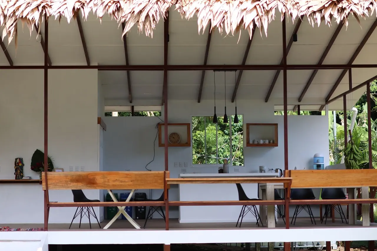 Espacio abierto con techo de palma, mesa larga de madera y cocina al fondo